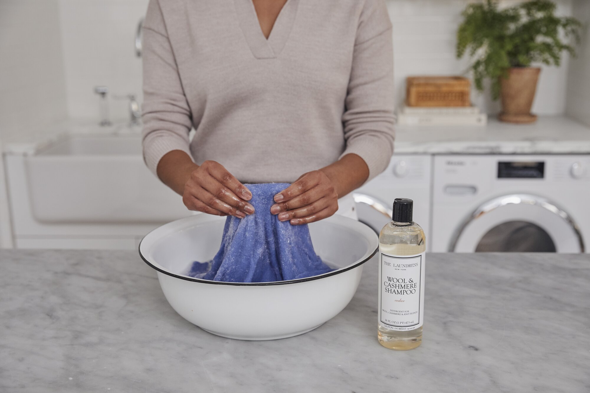 assets2f58e9d3cf0a1a474bb14df9285f98241d2f86e50cebb3cc4adca1eca9cada97fa94 A person washing a blue towel in a white basin utilizing The Laundress Wool & Cashmere Shampoo, surrounded by a clean and organized laundry space with plants.